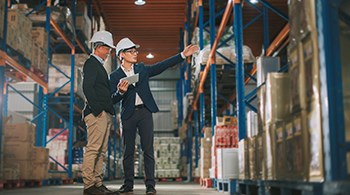 two-east-asian-men-in-white-hats-in-warehouse-one-with-tablet-in-one hand-gesturing-to shelf-with-other.jpg